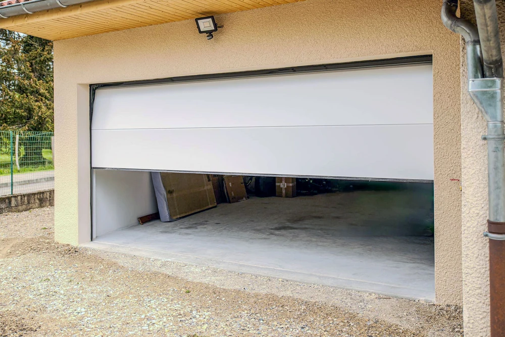 Garage résidentiel avec porte sectionnelle blanche partiellement ouverte, révélant l'intérieur vide avec sol béton et matériaux stockés