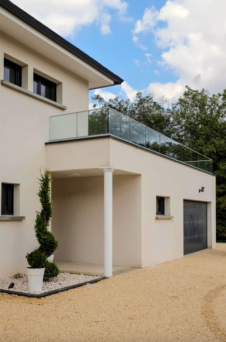 Maison contemporaine à deux niveaux avec façade en enduit beige clair, balcon avec garde-corps en verre, porche d'entrée soutenu par pilier blanc